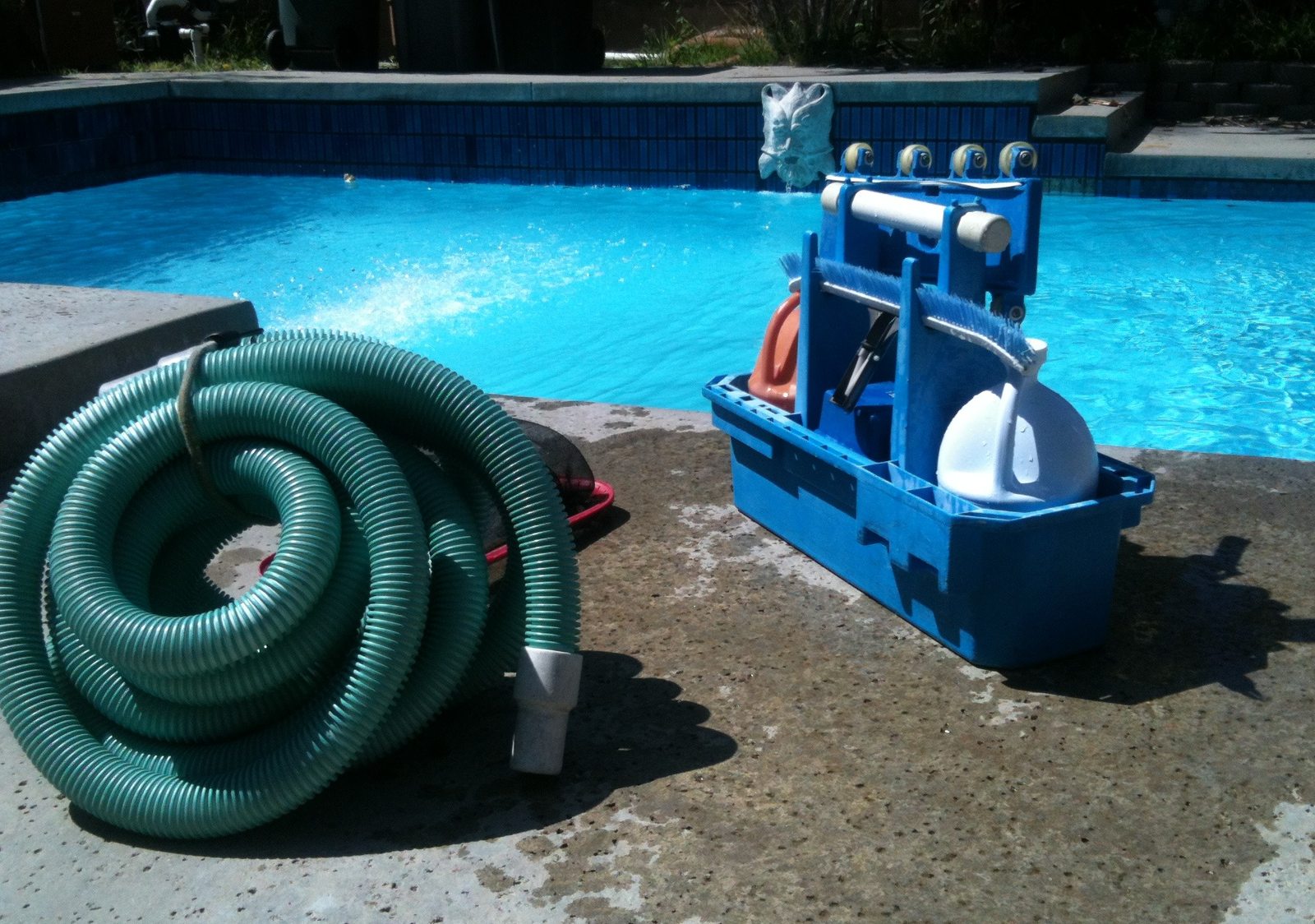 Pool vacuum hose, brushes, and a supply caddy on a concrete deck beside a tiled pool. Photo from the Clean Blue Pool site.